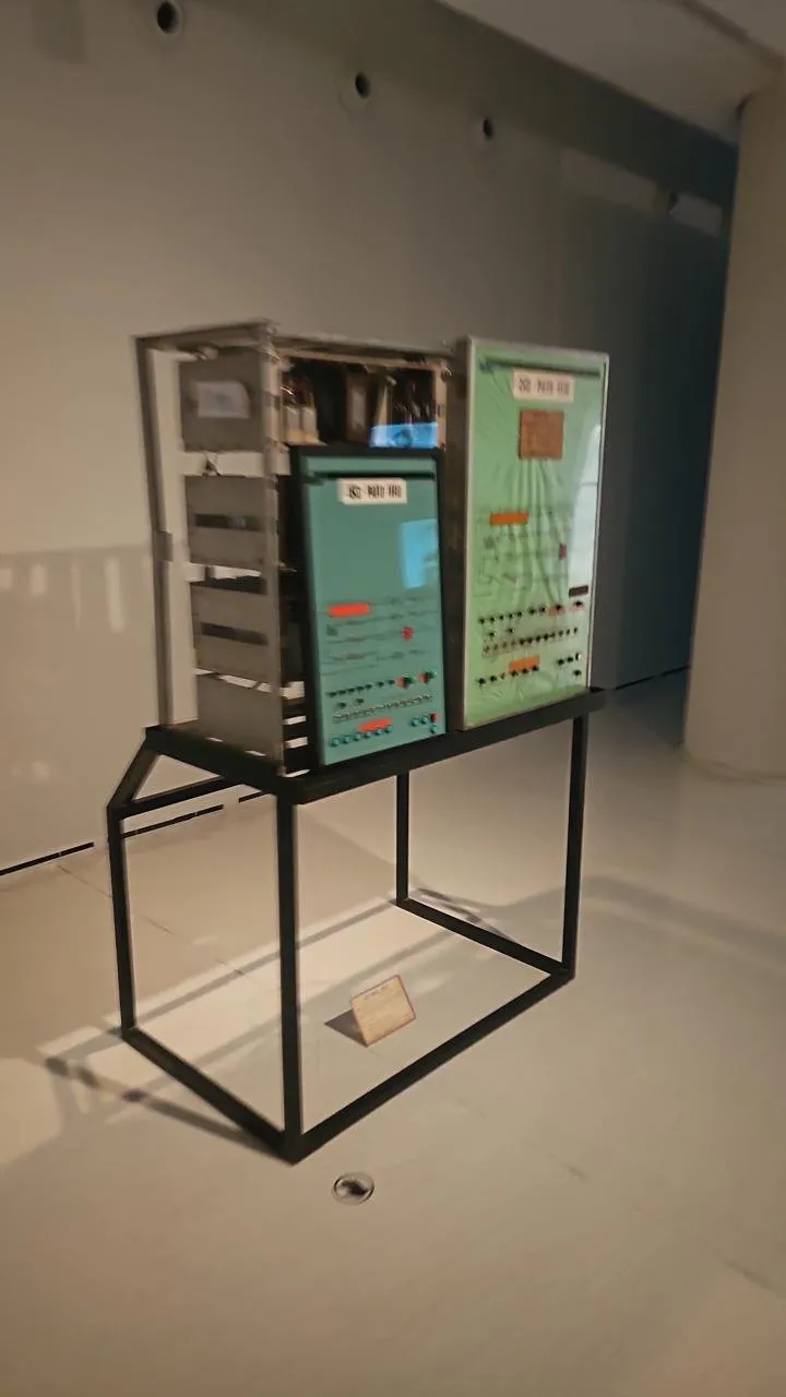 Peça instalada sobre suporte metálico aberto, com etiqueta de identificação no piso, em sala expositiva de piso claro